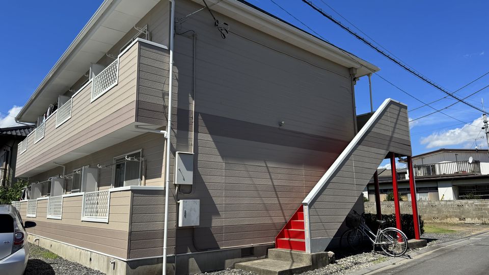 2 階建ての建物を表側から見た外観。2 階へ上がる外階段は赤色