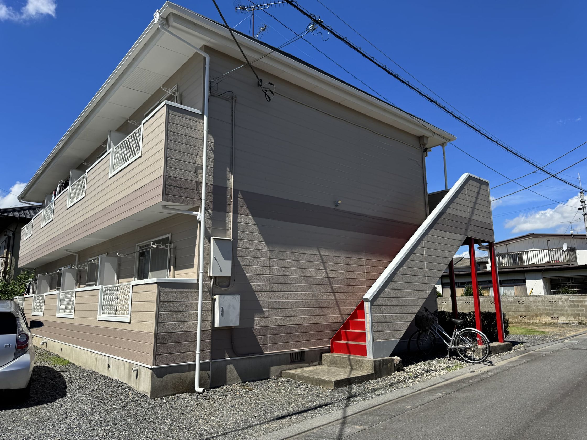 2 階建ての建物を表側から見た外観。2 階へ上がる外階段は赤色